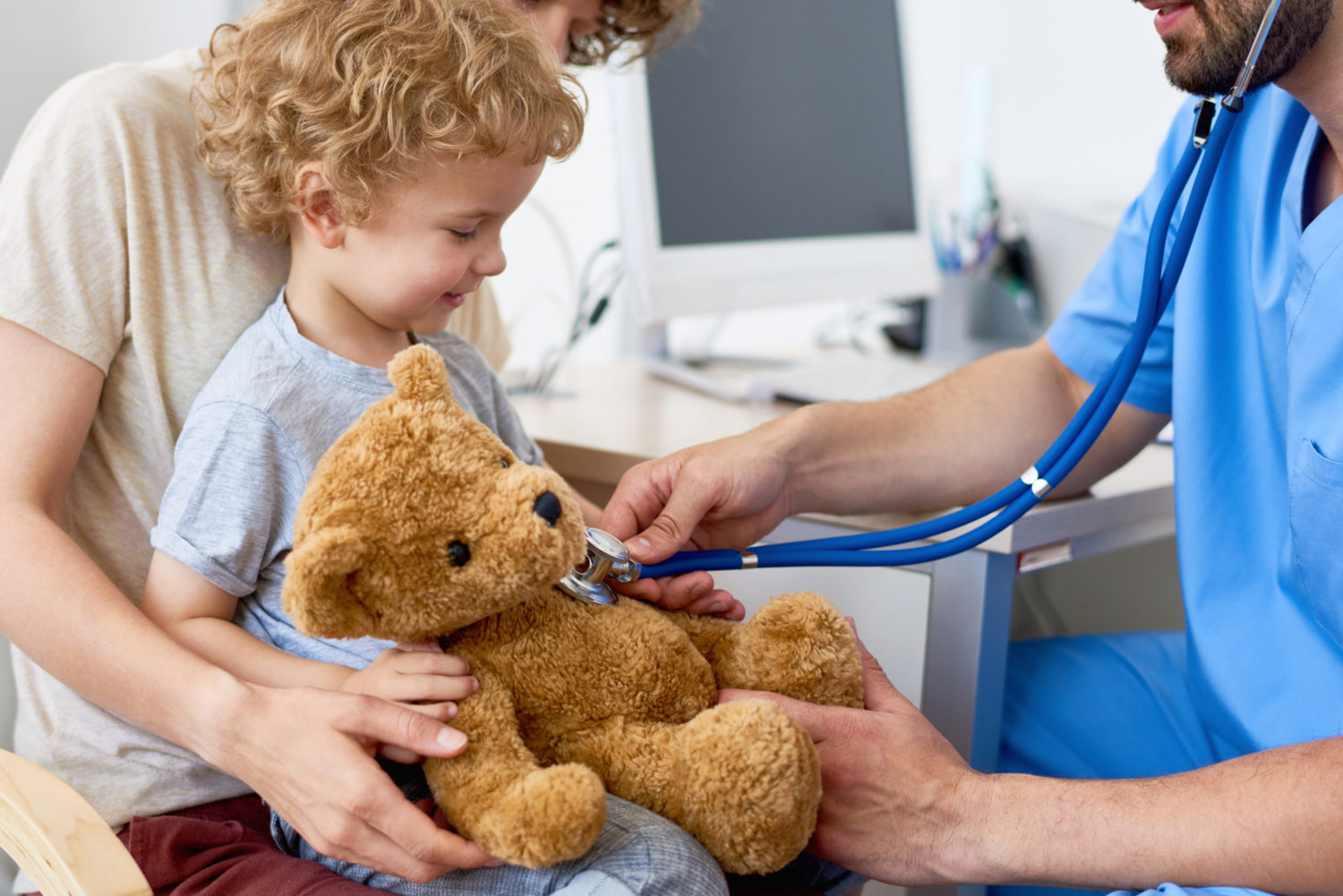 Portrait of adorable curly child  sitting on mothers lap in doctors office holding teddy bear toy, with pediatrician listening to heartbeat using stethoscope